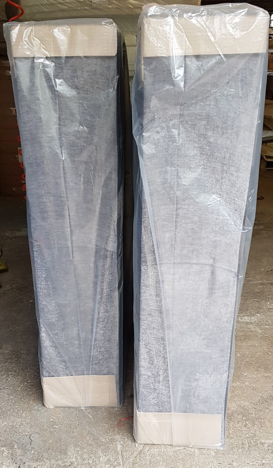 Two rectangular bed bases wrapped in protective plastic packaging, standing upright on a concrete floor.